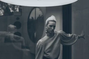 Monochrome fashion editorial portrait of a woman leaning against a glass wall with reflective surfaces.