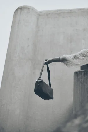 Arm of a model in a lace sleeve holding up a dark leather handbag against a concrete wall