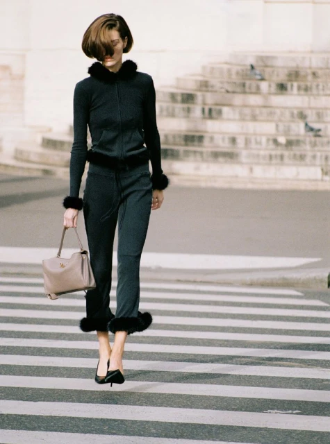 Woman in a black fur-trimmed tracksuit and heels walks across a crosswalk with a purse; architecture and pigeons in the background.