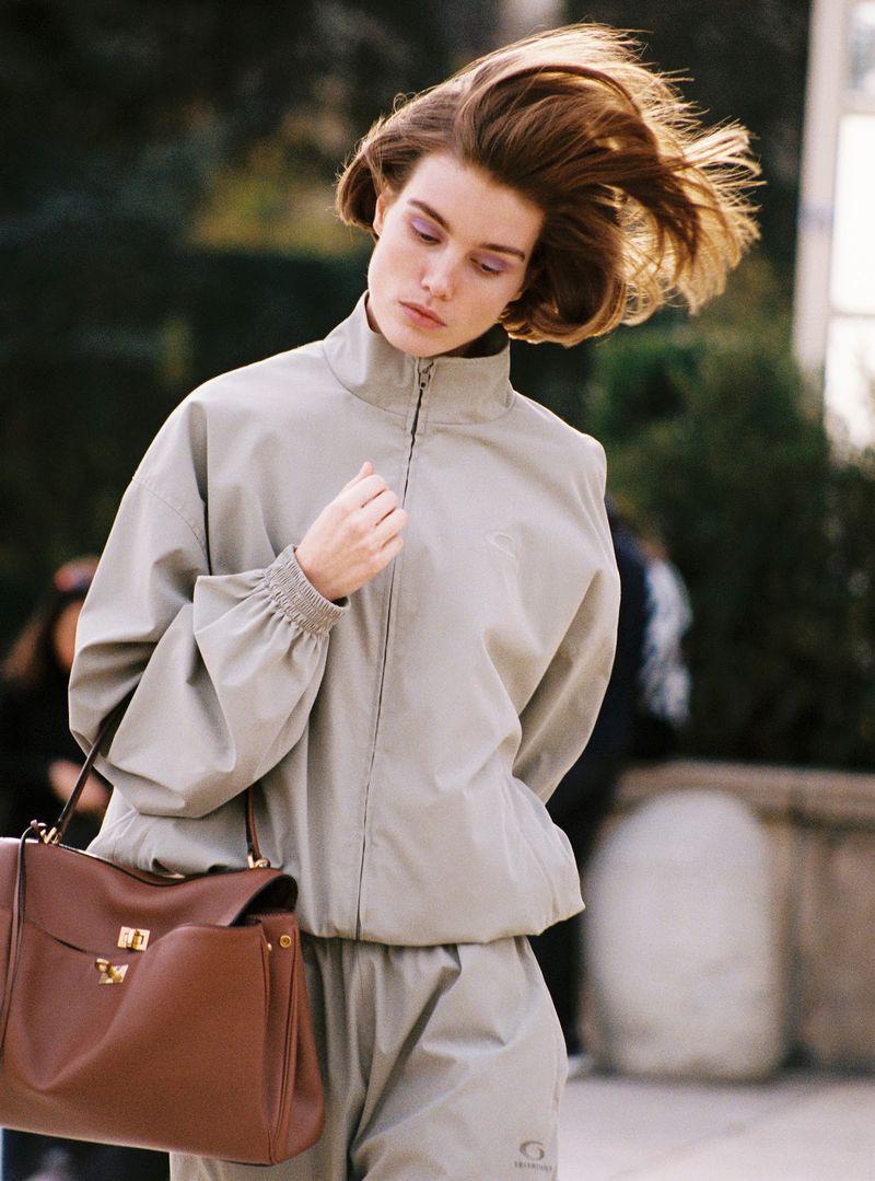 Woman in beige tracksuit with brown leather purse outdoors, captured in a medium shot with natural lighting and a blurred background.
