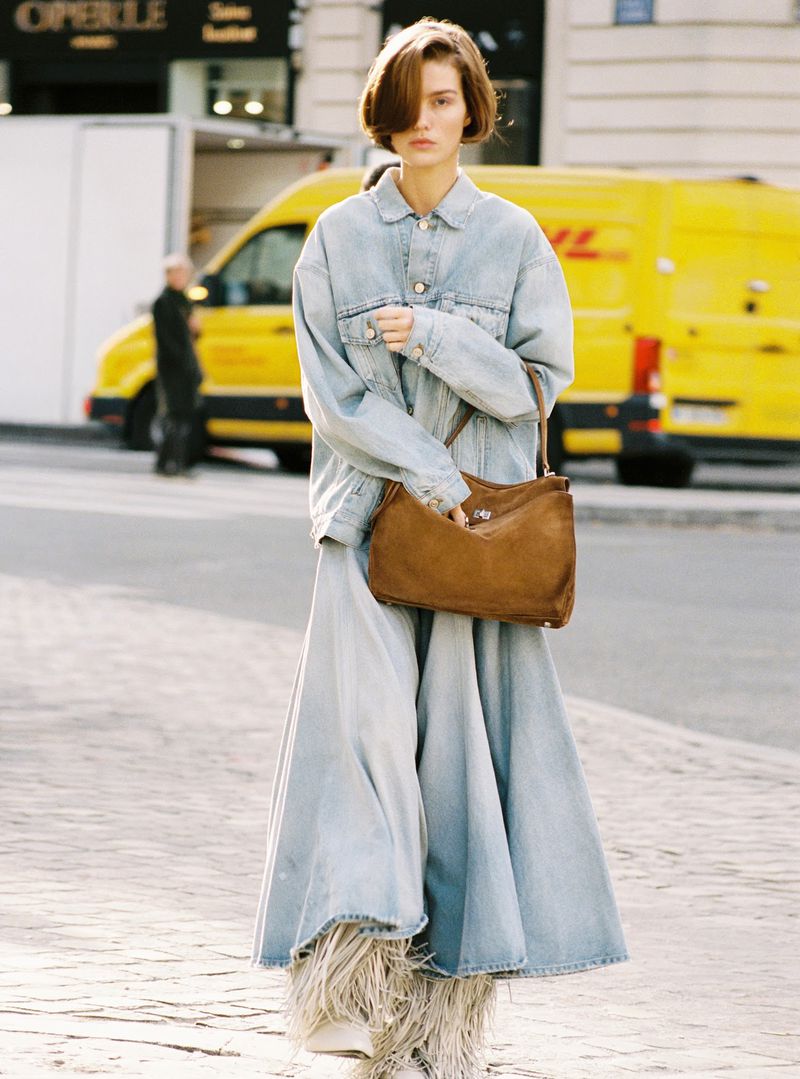 A woman wearing a denim jacket and long skirt walks on the street. She carries a brown bag on her shoulder and yellow van is parked nearby.