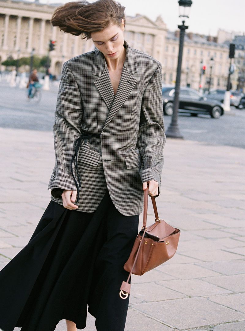 A woman in a houndstooth blazer and black skirt poses with a purse in an urban setting with Beaux-Arts architecture in the background.