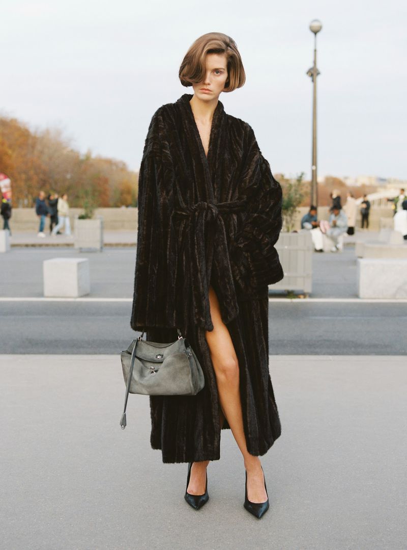 Elegant woman in a long dark brown fur coat stands on a city street carrying a grey leather handbag and wearing black heels.