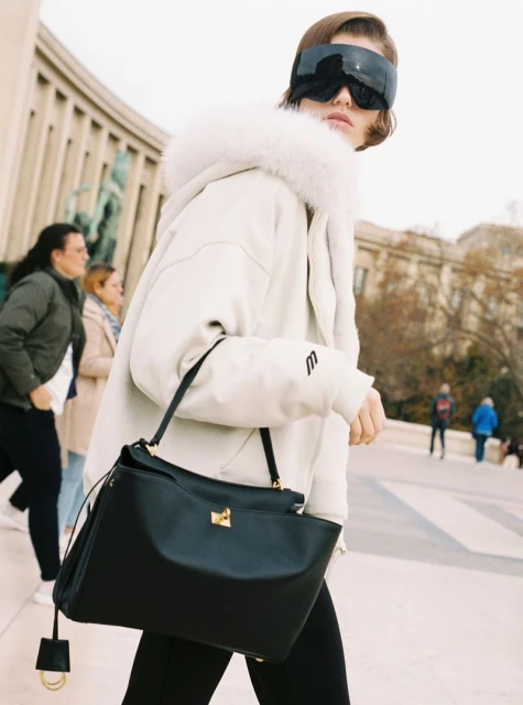 Woman in white hooded jacket, black leggings, and futuristic sunglasses carrying a black leather handbag outdoors.