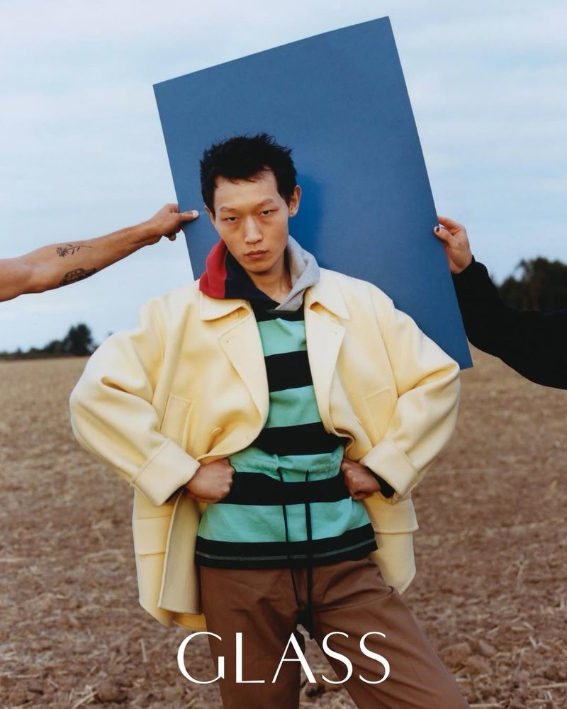 Editorial fashion photo of a young Asian man in a yellow jacket and striped shirt, flanked by disembodied arms holding a blue panel.