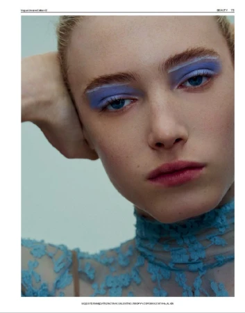 Close-up of a young blonde woman with bright blue eyeshadow, styled eyebrows, and a high-necked lace top, set against a light background.