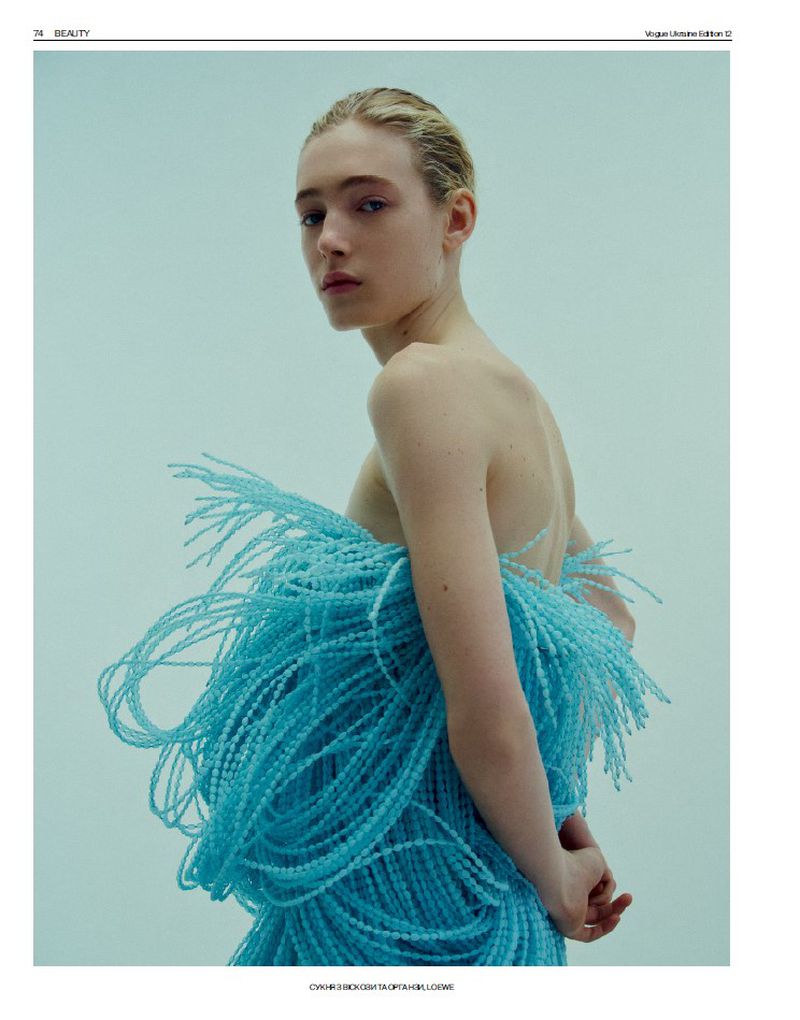 Blonde woman with bare shoulders poses in light blue beaded garment against a pale blue background in a beauty editorial shot.