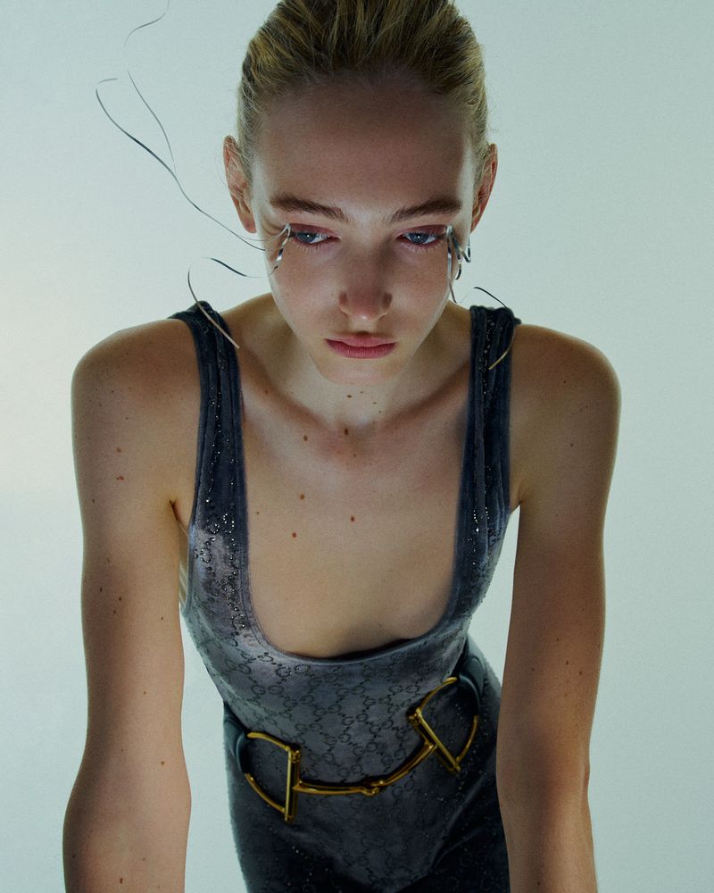 Young adult woman in a grey velvet bodysuit with a gold belt, standing in a brightly lit indoor space with soft, diffused lighting.