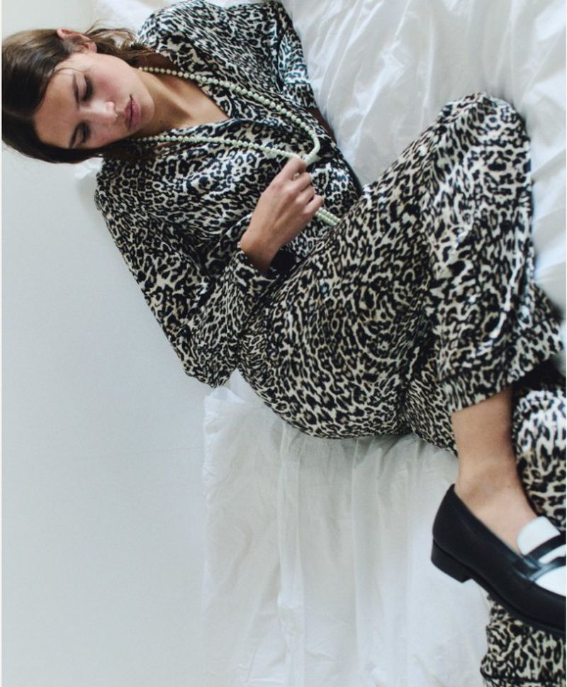 A brunette woman reclines on white sheets wearing an animal print pajama set and black and white loafers with a string of pearls.