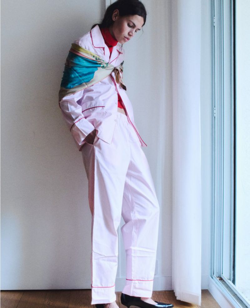 Fashionable woman in pink pajama-style suit by window with colorful scarf, black heels, and red top looking down.