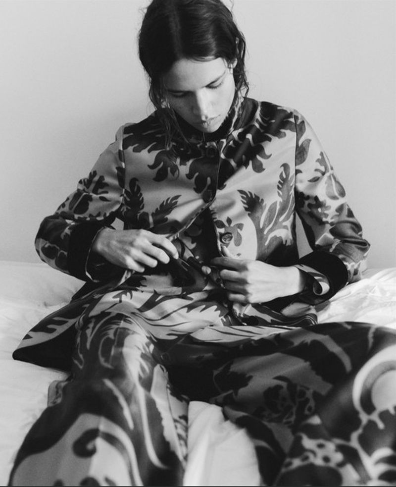 A woman sits on a bed wearing patterned loungewear and buttoning a matching jacket in a high-contrast indoor fashion shot.
