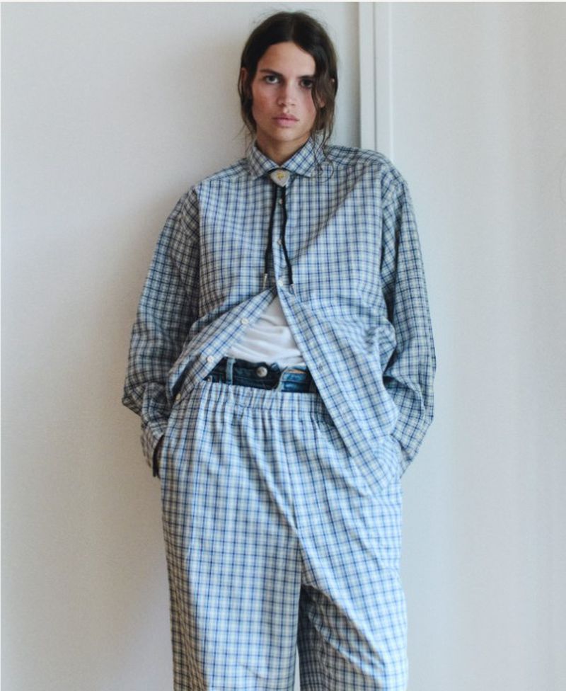 A person in a blue and white plaid outfit stands against a white wall, wearing a bolo tie in an indoor setting with natural lighting.