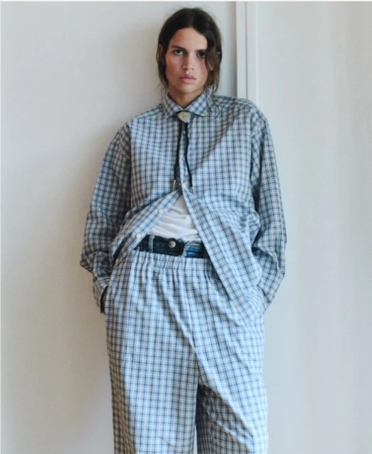 A person in a blue and white plaid outfit stands against a white wall, wearing a bolo tie in an indoor setting with natural lighting.