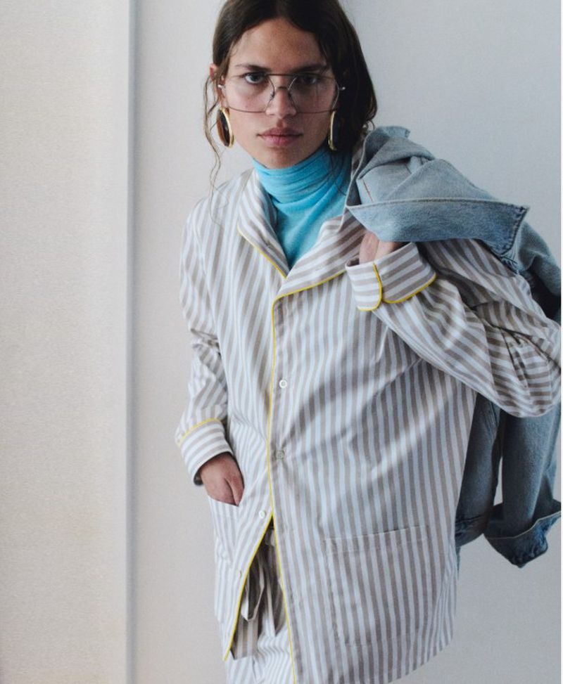 Model wearing wire-rim glasses, a striped pajama shirt over a blue turtleneck, and a denim jacket over one shoulder.