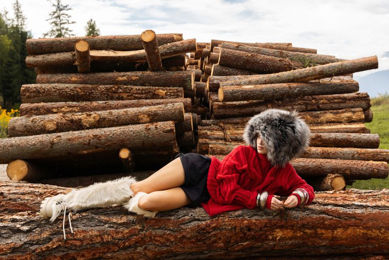 Woman in red sweater and fur hat lounges on a log pile in an outdoor fashion shot, trees and a cloudy sky in the background.