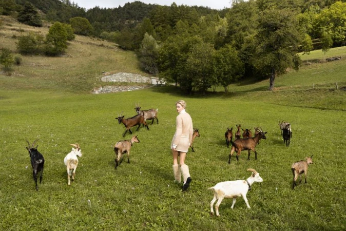 Woman in a light dress stands in a green meadow surrounded by a herd of goats, with rolling hills and trees in the background.