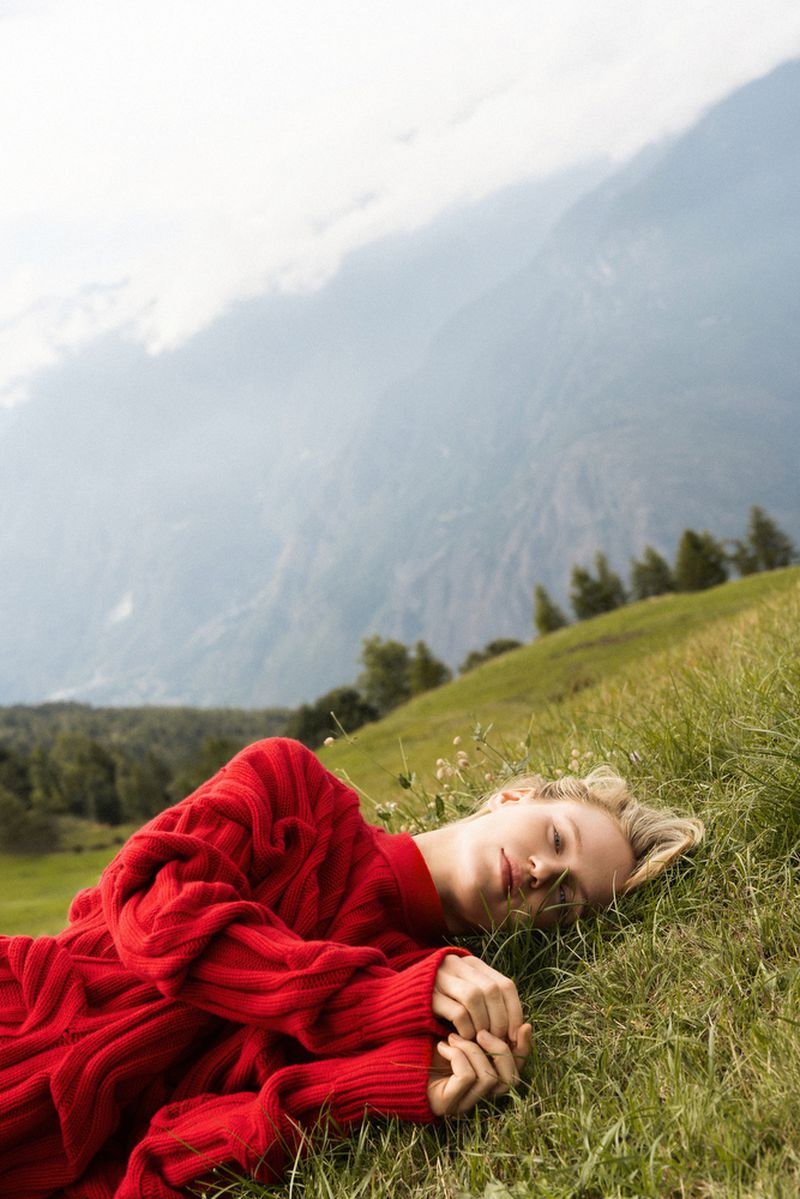 A woman with blonde hair lies on a grassy hillside wearing a red knit sweater, with a background of mountains and blue sky.