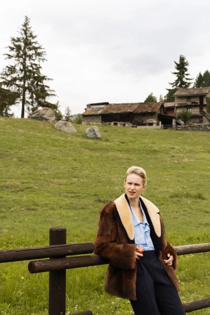 Adult female in fur coat leans on wooden fence, rural countryside setting with green grass, trees, and houses in the background.