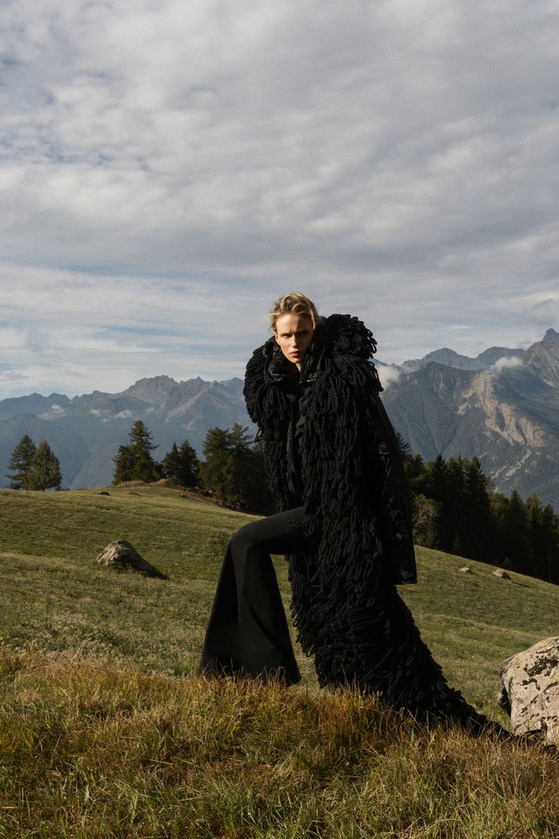 A woman poses in a black, fringed coat and flared pants in a mountain meadow beneath a cloudy sky, in a fashion editorial shot.