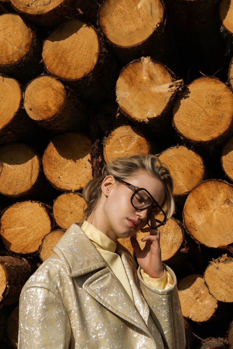 Woman in oversized glasses and a shimmery coat poses in front of a wall of stacked logs in natural lighting, fashion photography.