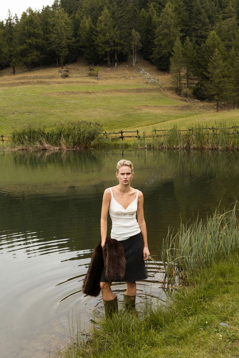 Blonde woman in white top, dark skirt, and boots stands in a lake holding a fur coat, with trees and green hills in the background.