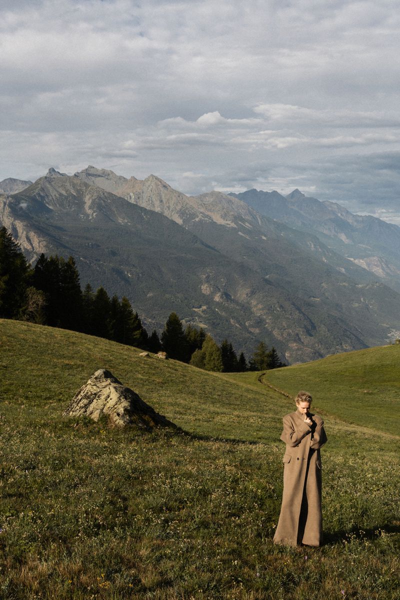Woman in a long coat standing in a green meadow with mountains in the background on an overcast day in a scenic outdoor fashion shot.