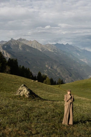Woman in a long coat standing in a green meadow with mountains in the background on an overcast day in a scenic outdoor fashion shot.