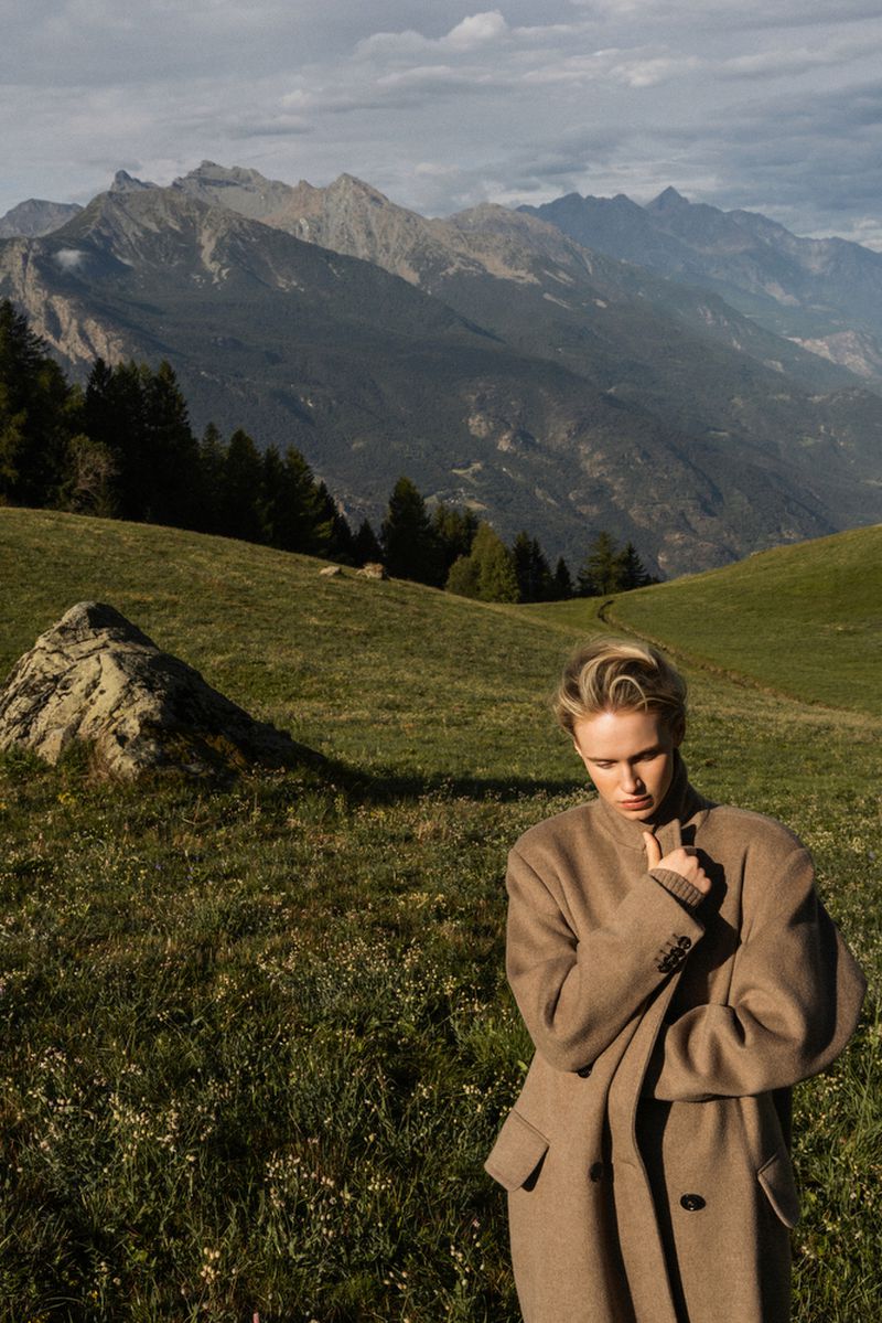 Woman in coat poses on grassy hillside with mountain backdrop on a cloudy day in outdoor fashion editorial photography.