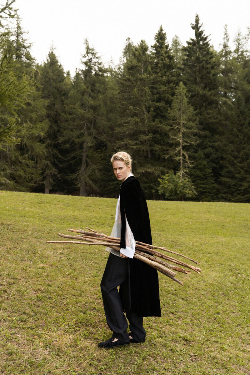 Woman in fashionable black cape carrying bundle of sticks on green grassy field, forest background. Outdoor setting.