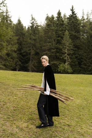 Woman in fashionable black cape carrying bundle of sticks on green grassy field, forest background. Outdoor setting.