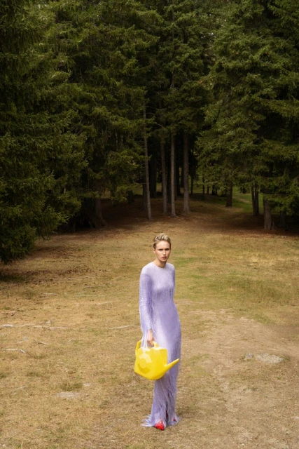 Young woman in a long lavender dress stands in a grassy field surrounded by tall trees, holding a yellow watering can.