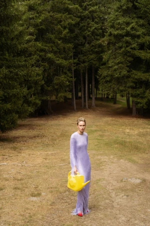 Young woman in a long lavender dress stands in a grassy field surrounded by tall trees, holding a yellow watering can.