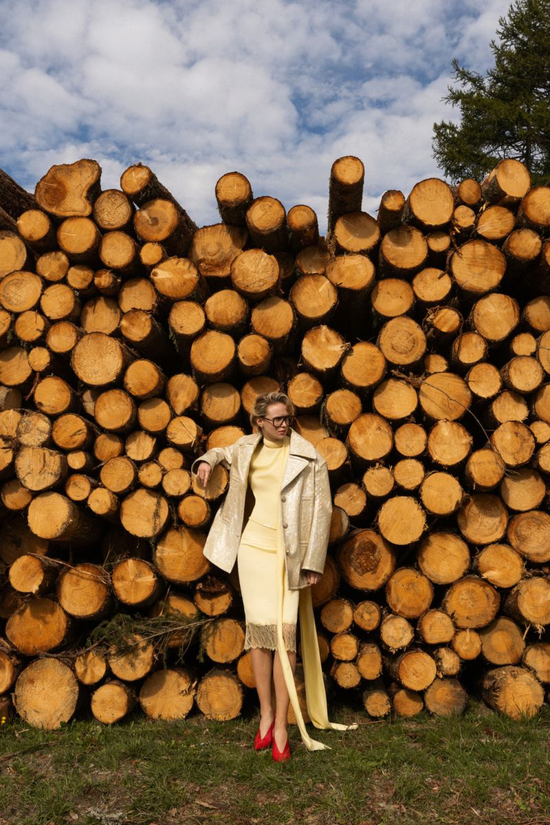 A woman in a yellow dress and beige jacket poses in front of a stack of cut logs, wearing red shoes against a blue sky backdrop.