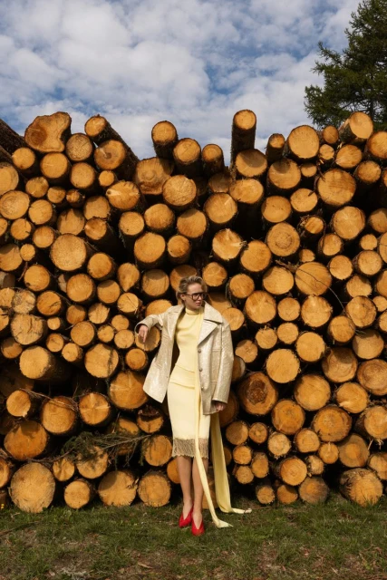 A woman in a yellow dress and beige jacket poses in front of a stack of cut logs, wearing red shoes against a blue sky backdrop.