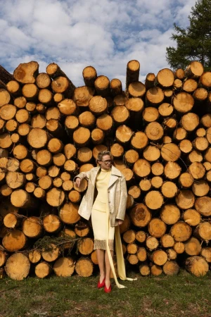 A woman in a yellow dress and beige jacket poses in front of a stack of cut logs, wearing red shoes against a blue sky backdrop.