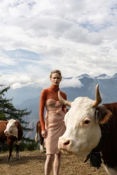 Model wearing a peach strapless dress and orange top standing in an alpine pasture with several cows.