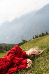 A blonde woman in an oversized red knit sweater lies on a grassy hillside in front of misty mountain peaks.