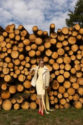Woman in pale yellow dress and metallic coat posing in front of a giant stack of logs under an open sky.