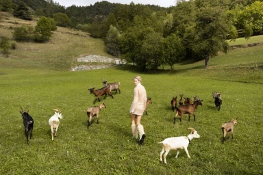 Model in a cream knit minidress and fuzzy boots walking through a grassy meadow surrounded by a herd of goats.
