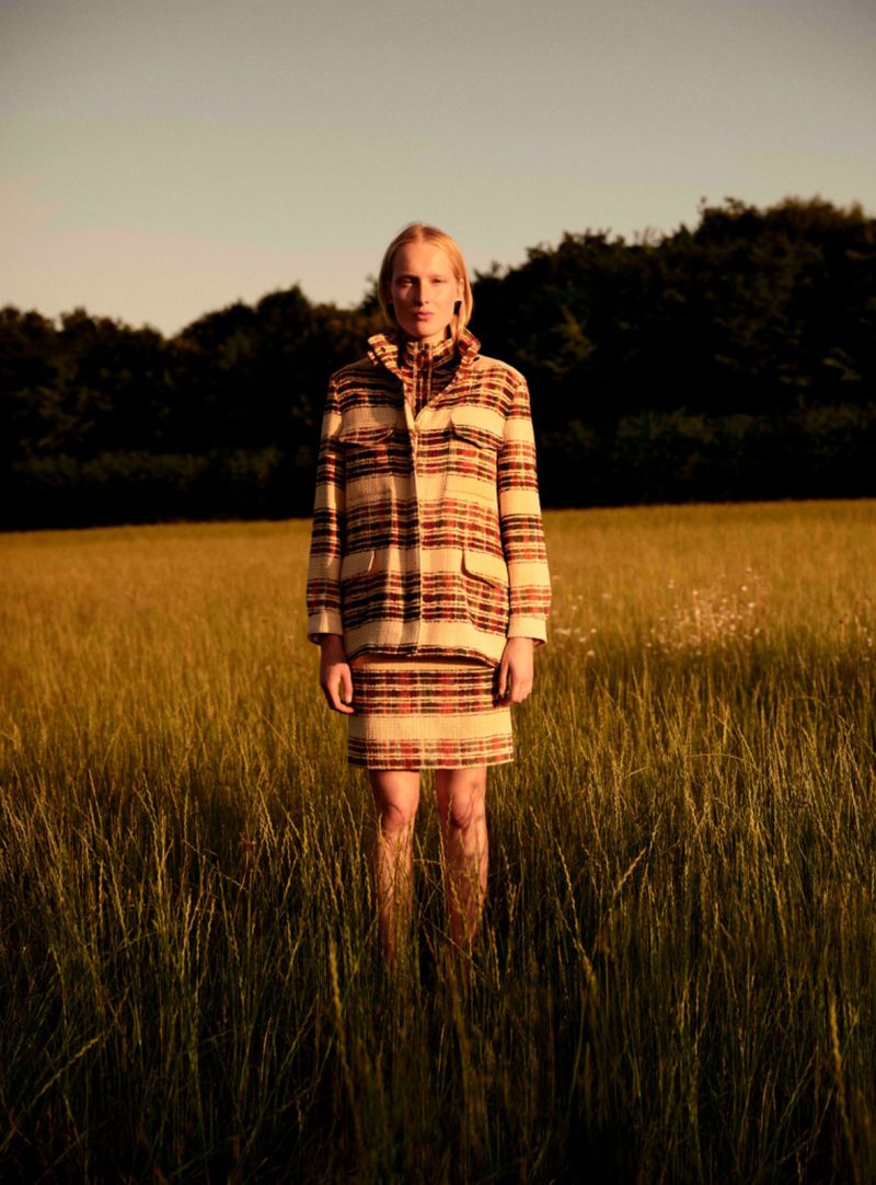 A woman in a striped coat and skirt stands in a field of tall grass against a backdrop of trees in the countryside at sunset.
