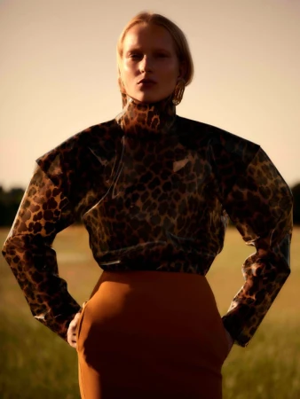 A fair-skinned woman in a leopard print top and brown skirt poses outdoors in a field, looking at the camera with a neutral expression.
