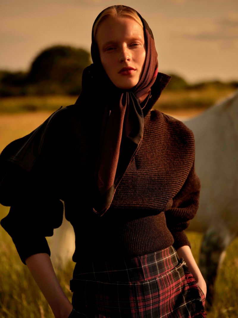 Young woman wearing a headscarf and a dark sweater in a rural setting, with a white horse visible in the background.