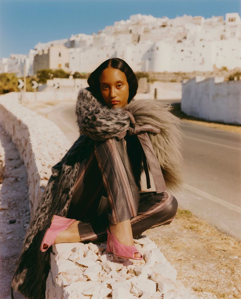 A woman with dark hair wearing a fur-trimmed jacket and pink shoes is sitting on a wall in front of white buildings and a road.