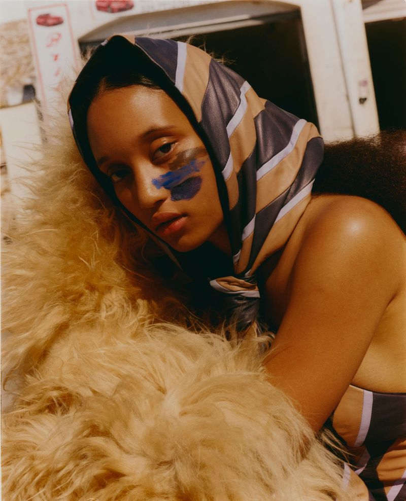 Portrait of a woman with face paint wearing a headscarf and striped top leaning against a light brown furry coat.