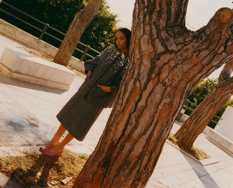 Outdoor fashion shoot of African-descent woman in coat and skirt, leaning against a tree in pink slippers during the daytime.