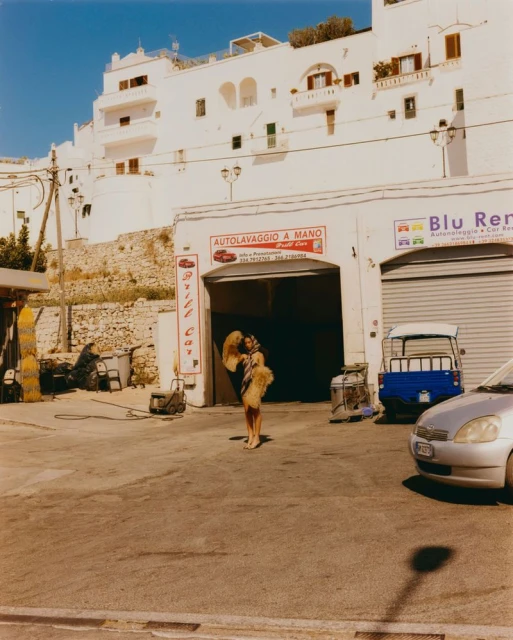 An adult female stands outside a car wash in front of white buildings on a sunny day with a silver car parked in foreground.