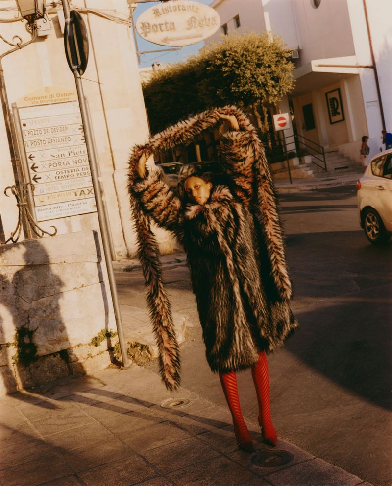 Fashionable adult woman poses on city sidewalk in oversized faux fur coat with red stockings in bright sunlight near buildings and street sign.