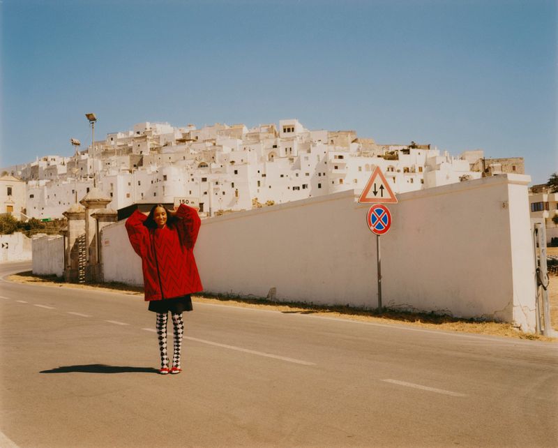 A young adult woman poses on a road in a red jacket, black and white patterned leggings, and red shoes, with a white walled city backdrop.