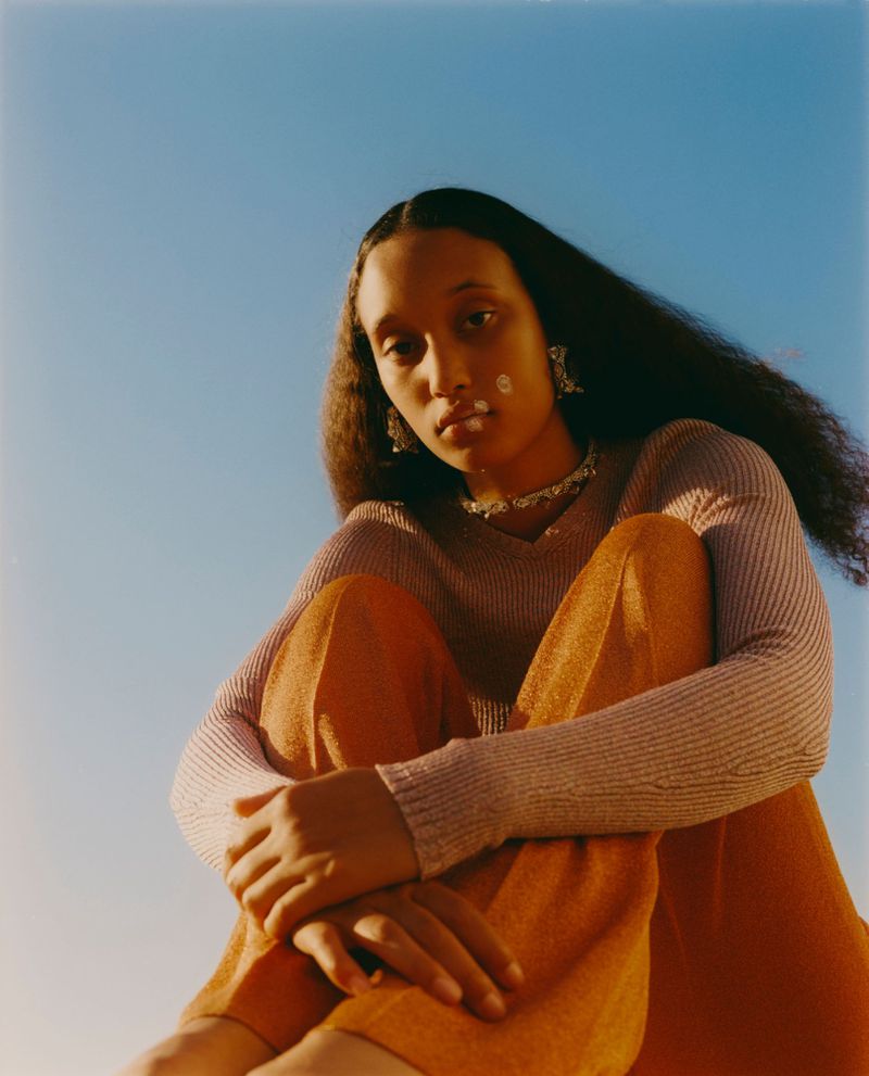 Young adult woman with long dark hair sits against a blue sky wearing orange pants and a lavender long-sleeve shirt with jewelry.