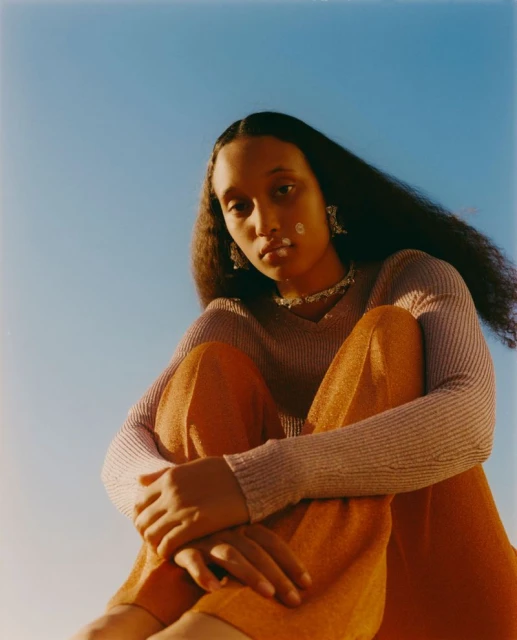 Young adult woman with long dark hair sits against a blue sky wearing orange pants and a lavender long-sleeve shirt with jewelry.